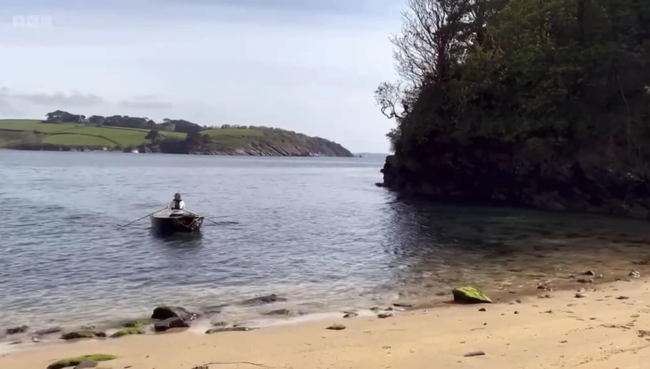 Sadie Jewellery rowing a small boat toward a hidden sea glass beach in Cornwall, surrounded by turquoise water and rugged coastline during filming.